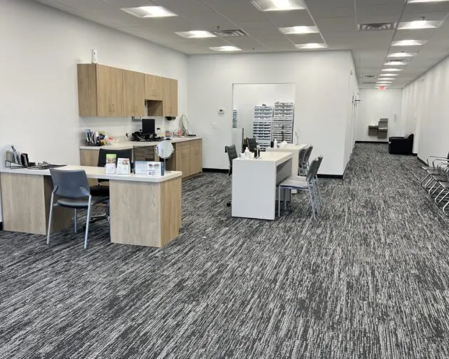 An eye care office with desks, chairs, and cabinets on a grey carpeted floor, and a corridor extending to the right.
