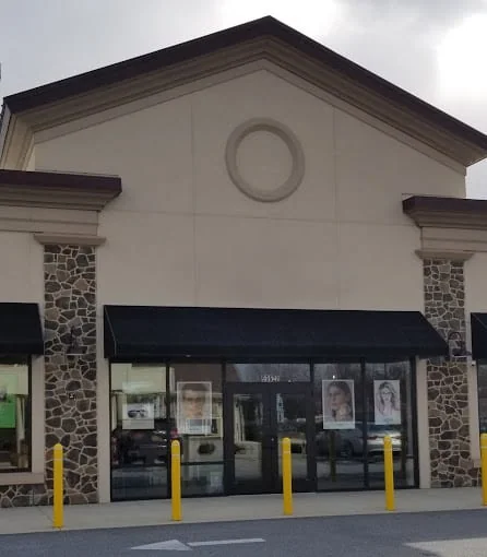 A beige stone-clad building with a pointed roof and black awnings. Posters with faces are displayed in the glass front windows, and there are yellow bollards in front.