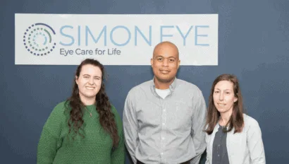 Three people standing in front of a sign that reads "Simon Eye: Eye Care for Life." They are all facing the camera and smiling.