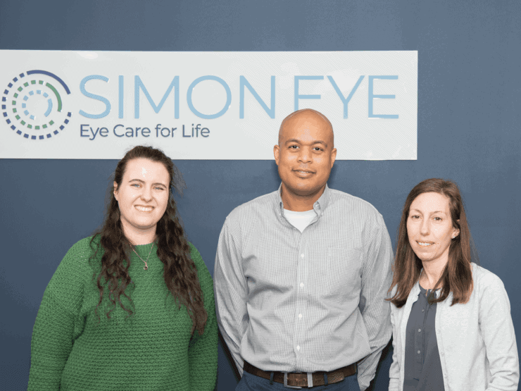 Three people standing in front of a sign that reads "Simon Eye: Eye Care for Life." They are all facing the camera and smiling.
