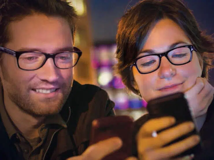 A man and woman, both wearing eyeglasses, look at their cellphones.