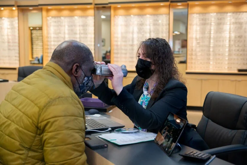 A woman sits across from a man at a desk, holding a tool up to his eyes to take a measurement.