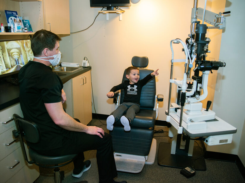 A young boy sitting in an eye exam chair points to eye exam equipment.