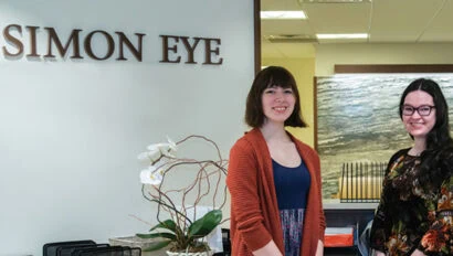 Two women standing in front of a sign that reads, "Simon Eye".