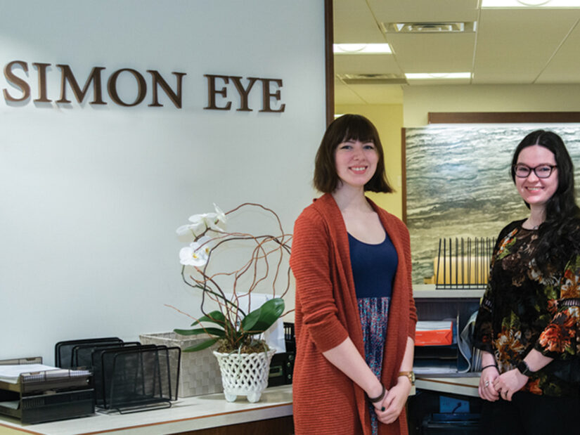 Two women standing in front of a sign that reads, "Simon Eye".