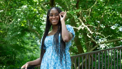 A person with long braided hair and wearing a blue floral dress stands outdoors by a railing, adjusting their glasses and smiling.