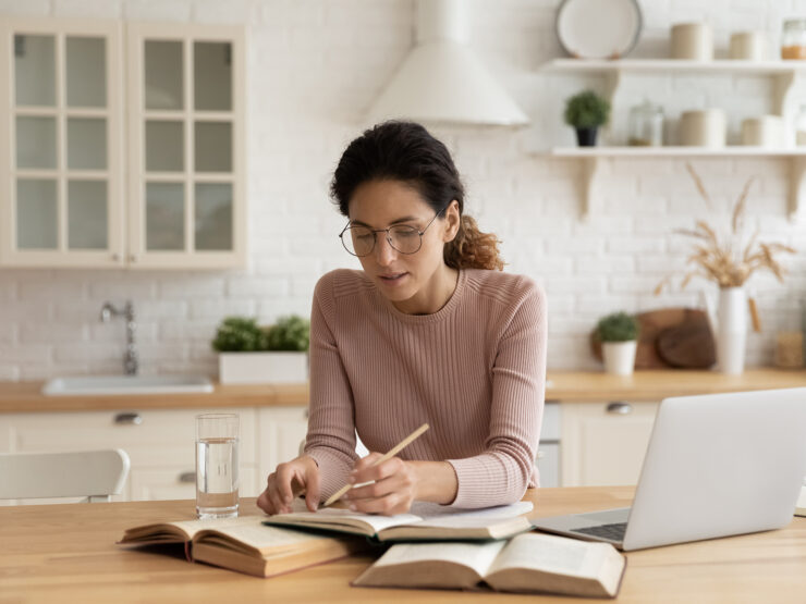 Woman in eyeglasses writing in a notebook next to her laptop.