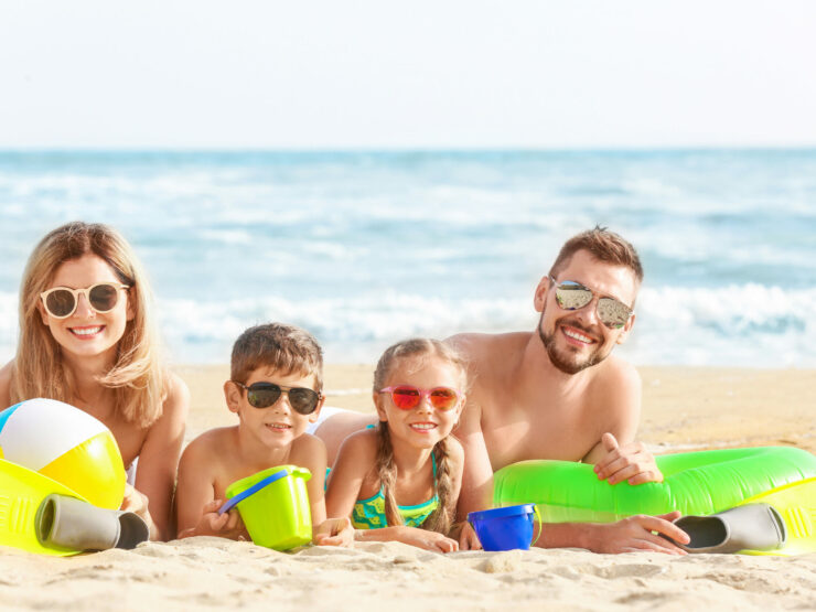 Family of four wearing sunglasses and lying on the sand at the beach.