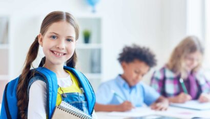 Smiling girl with rucksack and notepads with other school kids in the background.
