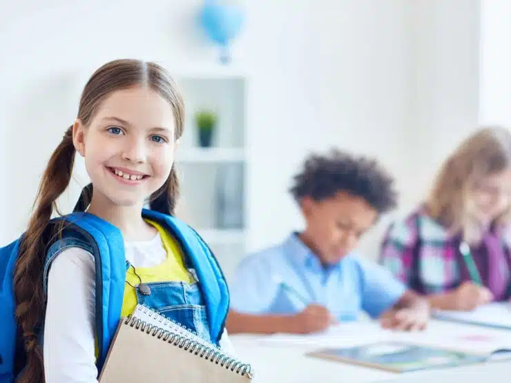 Smiling girl with rucksack and notepads with other school kids in the background.