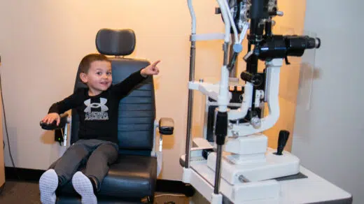 Boy sitting in a chair and pointing at a piece of eye equipment during a pediatric eye exam.
