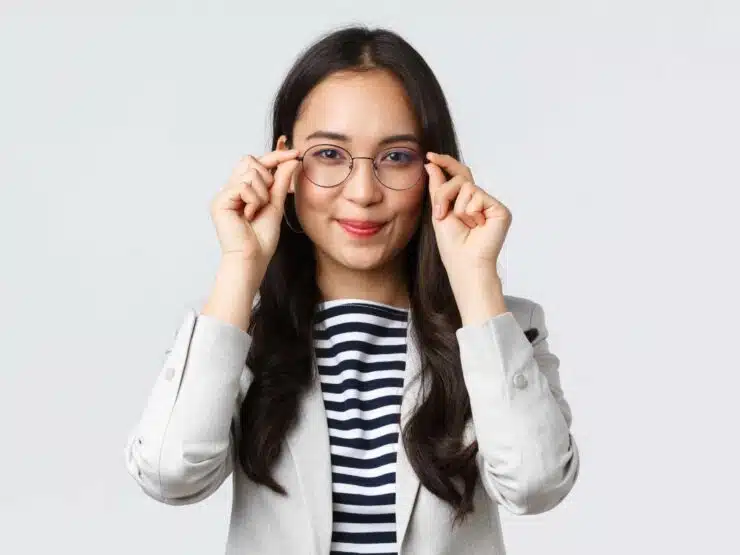 Woman in a grey jacket and striped shirt, smiling and putting on a pair of glasses.