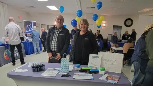 Two people stand behind a display table at an indoor event. The table has pamphlets, a gift bag, and other small items.