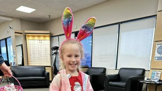A young child smiles while wearing rainbow-colored bunny ears in a waiting room with black chairs and large windows.