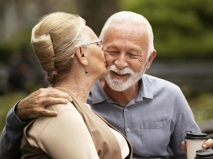 An elderly woman kisses an elderly man on the cheek as they smile. The man holds a takeaway coffee cup, and they are outdoors.