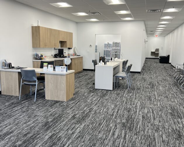 An eye care office with desks, chairs, and cabinets on a grey carpeted floor, and a corridor extending to the right.