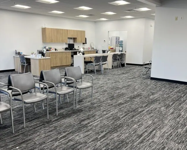 A large, clean office space with multiple chairs arranged in rows, a kitchenette with wooden cabinets, and a few tables and chairs near the back wall.