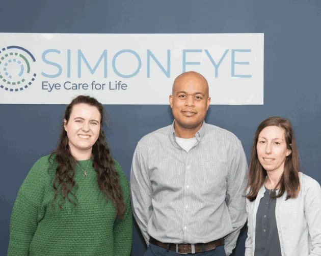Three people standing in front of a sign that reads "Simon Eye: Eye Care for Life." They are all facing the camera and smiling.