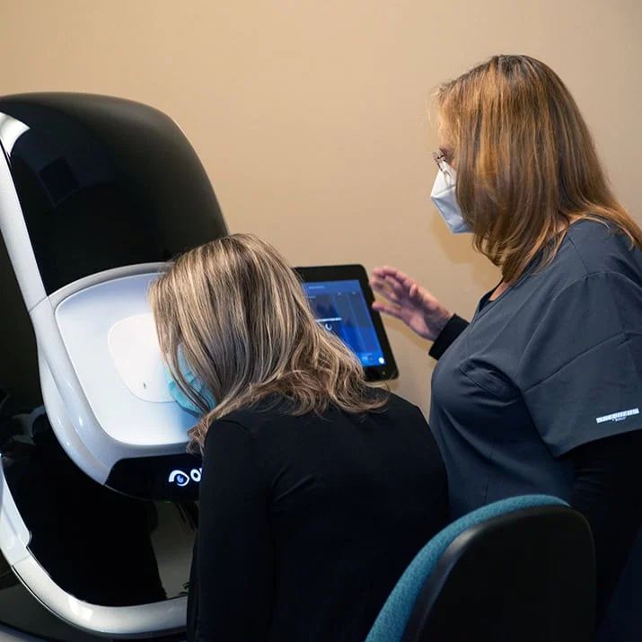 A woman checks her eye at machine while a doctor stands beside her.