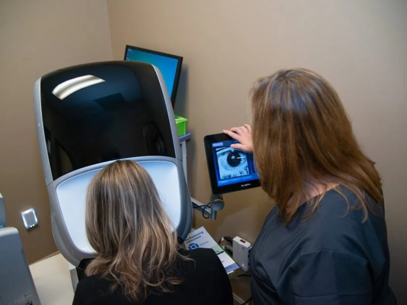 A patient leaning her head on eye exam equipment while a doctor monitors a screen.