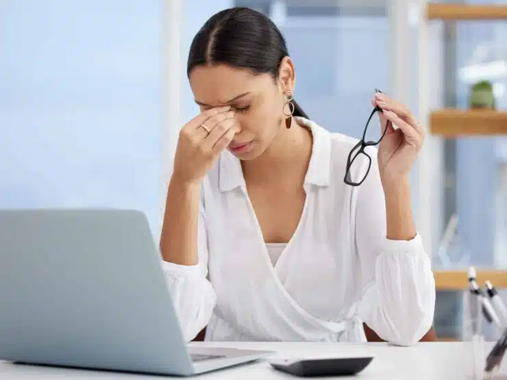 A person in a white shirt is sitting at a desk, holding glasses and rubbing the bridge of their nose, appearing stressed or tired.