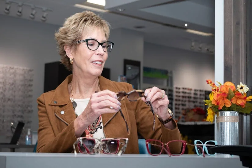 A woman in a brown jacket is examining a pair of sunglasses in an eyewear store, with several other pairs of glasses displayed in front of her.
