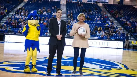 Two people stand on a basketball court, one holding a basketball. A person in a bird mascot costume stands beside them.