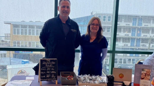 Two people stand behind a booth with various items displayed, including a board with "20/20/20 Rule" written on it and a container of small objects.